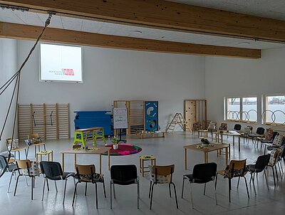 Ein großer, hell gestalteter Raum, der normalerweise als Turnraum genutzt wird. Darin stehen Stühle, die im Kreis angeordnet sind und drei kleinere Tische. Auf jedem der Stühle liegt ein buntes Kissen. An den Wänden befinden sich Regale mit Spiel- und Sportgeräten wie einer Holzleiter, blauen gestapelten Turnmatten und einem kleinen Klettergerüst. Im Hintergrund sind ein Flipchart mit Notizen und ein runder roter Teppich zu sehen, auf dem in der Mitte ein Pflanze platziert ist. Links oben befindet sich ein großes Fenster mit Milchglas, auf dem spiegelverkehrt das Logo der ESO Education Group zu erkennen ist.