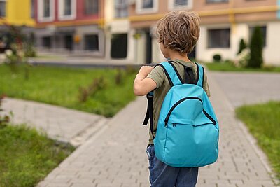 Rückansicht eines kleinen Junge mit einem blauen Rücksack, der zu einem Kindergarten geht. Im Hintergrund sind gepflasterte Wege, Grasflächen und bunte Gebäude zu sehen.