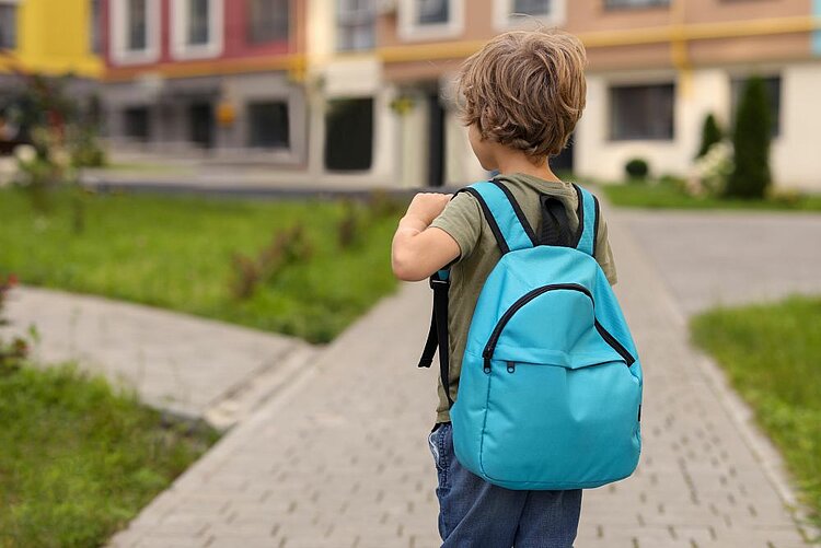 Rückansicht eines kleinen Junge mit einem blauen Rücksack, der zu einem Kindergarten geht. Im Hintergrund sind gepflasterte Wege, Grasflächen und bunte Gebäude zu sehen.