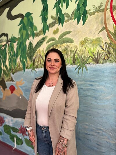 Interview mit unserer Leiterin Verwaltung, Kundenbetreuung und Personal KITA Aufnahme von Jeanine Grünheid vor einer bunt bemalten Wand.