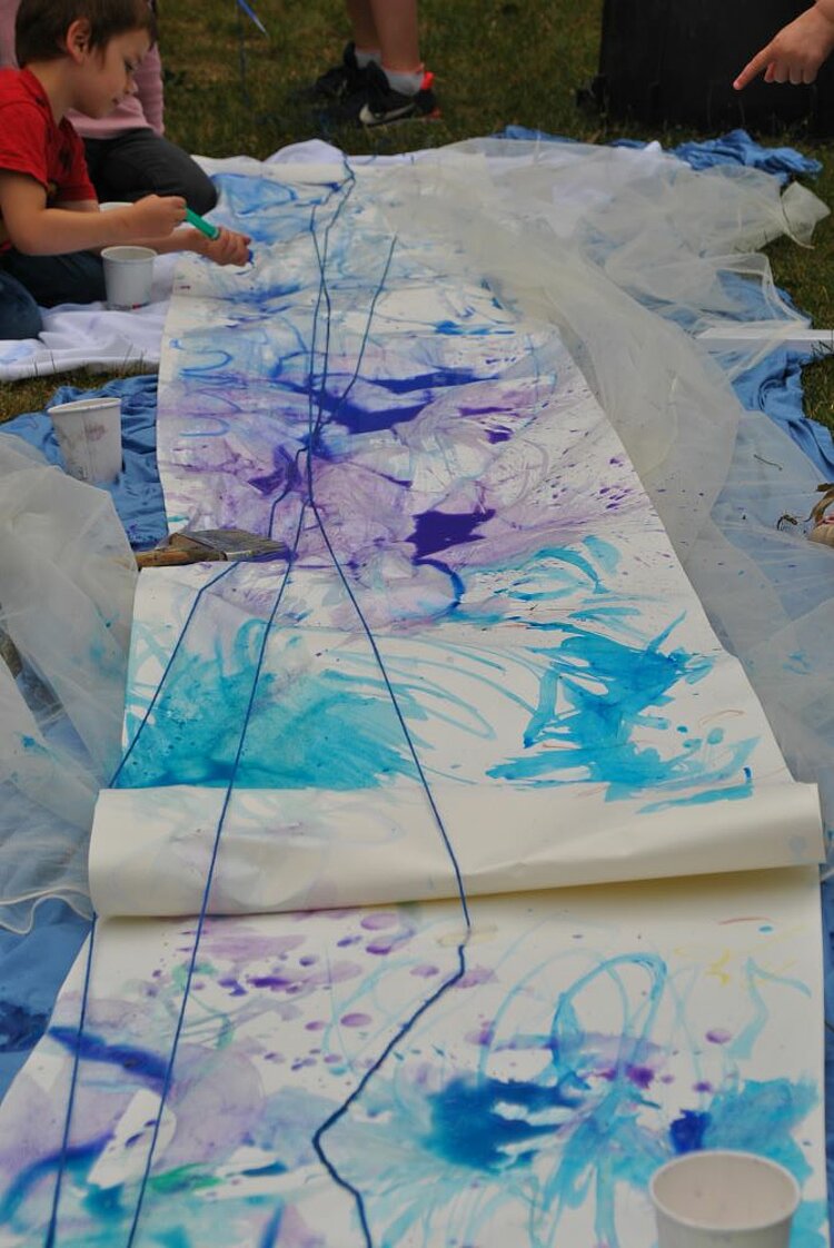 Sommerfest mit Spiel und Spaß Ein Kind malt mit Pinsel und Wasserfarbe auf ein langes Papier auf dem Boden. Die bunten Blau- und Violetttöne verlaufen ineinander – Teil des Wasserprojekts beim Sommerfest.