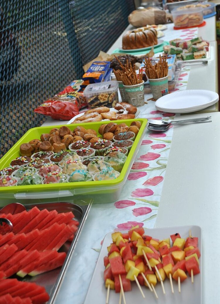Sommerfest mit Spiel und Spaß Ein langes Buffet aus mitgebrachten Speisen, wie Käsespieße, Melone, Muffins und Kuchen.