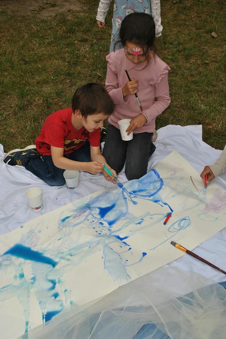 Sommerfest mit Spiel und Spaß Zwei Kinder malen mit Pinsel und Wasserfarbe auf ein langes Papier auf dem Boden. Die Blautöne verlaufen ineinander – Teil des Wasserprojekts beim Sommerfest.