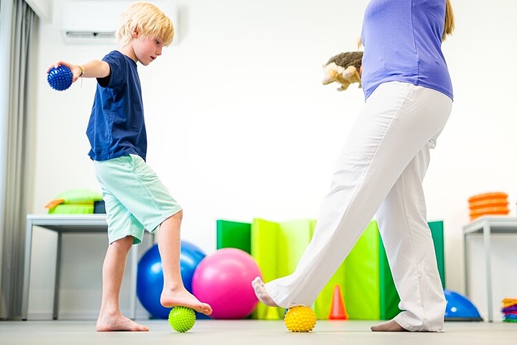 Ein barfuß stehendes Kind balanciert in einem hellen Therapieraum vorsichtig auf einem gelben Igelball, während es in der Hand einen blauen Massageball hält. Eine erwachsene Fachkraft, vermutlich eine Ergotherapeutin oder pädagogische Betreuungsperson, unterstützt die Übung und steht stabil daneben. Im Hintergrund befinden sich bunte Gymnastikbälle, Schaumstoffelemente und Spielmaterialien. Die Szene zeigt eine spielerische Bewegungs- und Gleichgewichtsübung zur Förderung der Motorik, Körperwahrnehmung und sensorischen Integration bei Kindern. Das Bild steht für frühkindliche Förderung, Ergotherapie, Physiotherapie und bewegungsorientierte Pädagogik in einem professionellen, kindgerechten Umfeld.