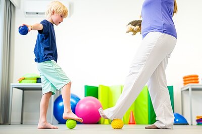 Ein barfuß stehendes Kind balanciert in einem hellen Therapieraum vorsichtig auf einem gelben Igelball, während es in der Hand einen blauen Massageball hält. Eine erwachsene Fachkraft, vermutlich eine Ergotherapeutin oder pädagogische Betreuungsperson, unterstützt die Übung und steht stabil daneben. Im Hintergrund befinden sich bunte Gymnastikbälle, Schaumstoffelemente und Spielmaterialien. Die Szene zeigt eine spielerische Bewegungs- und Gleichgewichtsübung zur Förderung der Motorik, Körperwahrnehmung und sensorischen Integration bei Kindern. Das Bild steht für frühkindliche Förderung, Ergotherapie, Physiotherapie und bewegungsorientierte Pädagogik in einem professionellen, kindgerechten Umfeld.