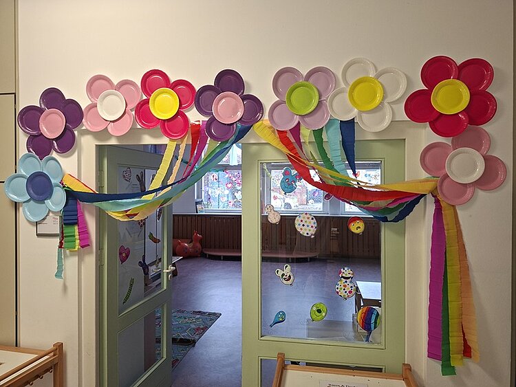 Bunte Papierblumen aus Papptellern sind oberhalb einer Tür zum Gruppenraum der Kita Waldräuber angebracht. Dazwischen verlaufen mehrere Girlanden aus farbigem Krepppapier in den Farben Blau, Gelb, Grün, Orange und Rot. An der Glastür und den Fenstern darunter sind weitere bunte Papierdekorationen befestigt. Im dahinterliegenden Raum ist ein Teppich mit buntem Muster und ein rotes Spielpferd zu sehen.