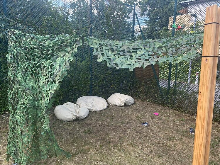 Ein schattiger Bereich im Garten, abgedeckt mit einem Tarnnetz, unter dem drei große weiße Sitzkissen auf dem Boden liegen. Im Hintergrund sind ein Zaun und Bäume zu sehen.
