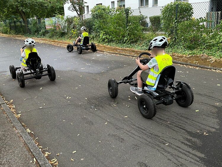 Drei Kinder in gelben Sicherheitswesten und Helmen fahren auf kleinen Go-Karts auf einem asphaltierten Weg in einem parkähnlichen Gelände.