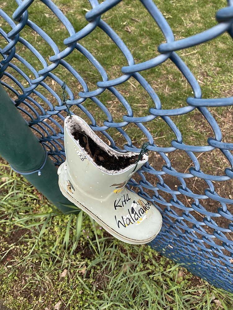 Ein einzelner, hellgrüner Gummistiefel hängt mit Draht an einem blauen Maschendrahtzaun. Im Inneren des Stiefels befindet sich Erde. Auf dem Stiefel steht handschriftlich der Text 'Kids Waldgarten'. Im Hintergrund ist eine Rasenfläche zu sehen.