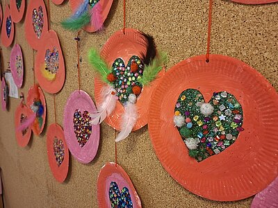 Mehrere runde Bastelarbeiten von Kindern der KITA Waldräuber aus bemaltem Papier sind an einer Korkwand befestigt. Jede Scheibe ist in kräftigen Rot- und Rosatönen gehalten und hat in der Mitte ein ausgeschnittenes Herz, das mit bunten Glitzersteinen, Pailletten und kleinen Federn verziert ist. Die einzelnen Elemente hängen an roten Fäden und sind unterschiedlich gestaltet. 