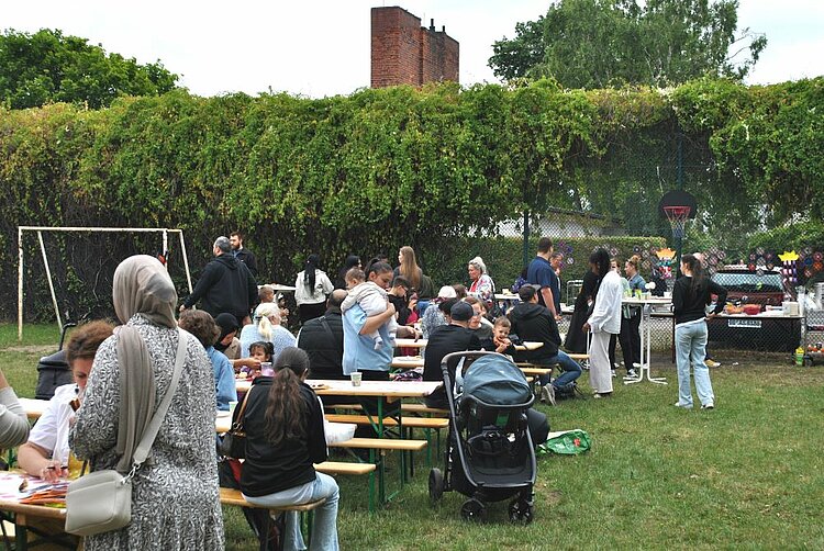 Sommerfest mit Spiel und Spaß Eltern, Kinder und Mitarbeitende sitzen und stehen an Bierzeltgarnituren auf einer Wiese. Im Hintergrund ist das Buffet aufgebaut, Menschen unterhalten sich in entspannter Atmosphäre.