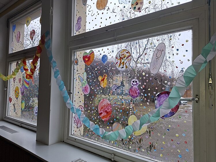 Bunte Papiergirlanden hängen vor großen Fenstern in der KITA Waldräuber. Die Fensterscheiben sind mit zahlreichen, handgemalten Bildern und Punkten aus Papier oder Folie dekoriert. Die Motive zeigen Herzen, Tiere und verschiedene Fantasiefiguren in kräftigen Farben. Draußen sind kahle Bäume und ein grauer Himmel zu sehen.