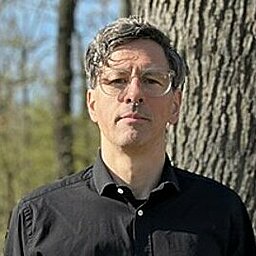 Mann steht in einem Waldgebiet vor einem dicken Baum. Er trägt eine schwarze Brille und ein schwarzes Hemd. Der Hintergrund ist lichtdurchflutet mit kahlen Bäumen.