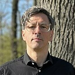 Mann steht in einem Waldgebiet vor einem dicken Baum. Er trÀgt eine schwarze Brille und ein schwarzes Hemd. Der Hintergrund ist lichtdurchflutet mit kahlen BÀumen.