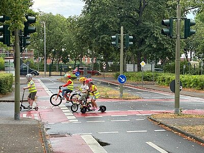 Eine Gruppe von Kindern in gelben Sicherheitswesten und Helmen überquert eine Kreuzung mit Ampeln mit Fahrrädern und Rollern. Im Hintergrund sind Bäume, Sträucher, ein Zaun und parkende Autos zu erkennen.