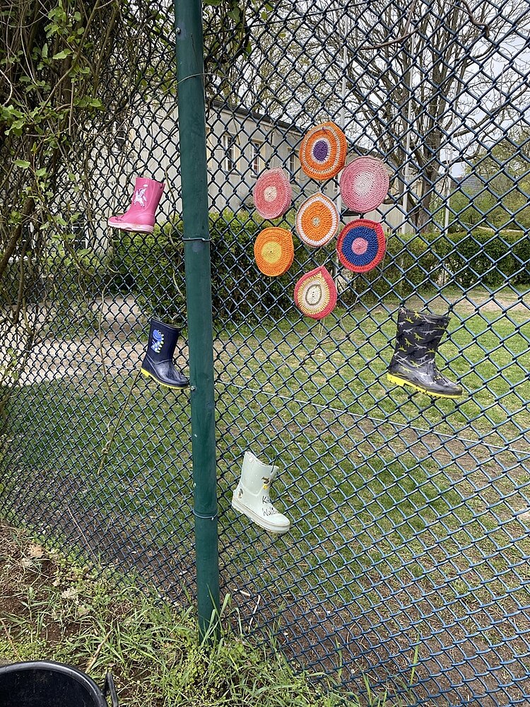 An einem Maschendrahtzaun hängen mehrere bunte Kindergummistiefel und gehäkelte runde Untersetzer in verschiedenen Farben. Im Hintergrund ist eine Rasenfläche und das Gebäude der KITA Waldräuber zu sehen. 