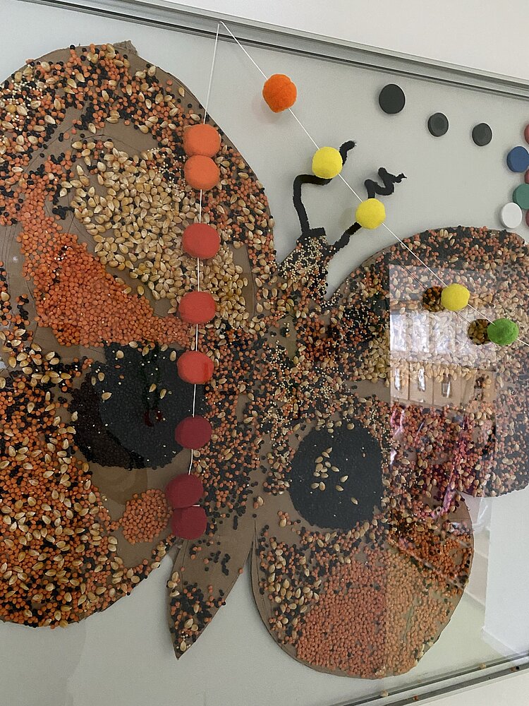 Große Schmetterlingscollage aus Körnern, Samen und bunten Pompons an einer Glasfläche.