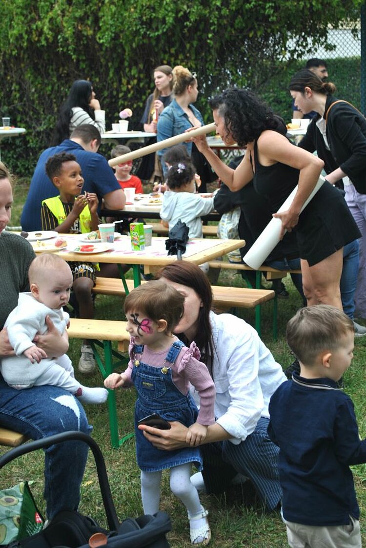 Sommerfest mit Spiel und Spaß Auf dem Bild ist eine lebhafte Szene eines Sommerfestes auf dem Außengelände einer KITA zu sehen. Mehrere Kinder und Erwachsene sind an langen Holztischen versammelt, essen und unterhalten sich. Ein kleines Mädchen mit Schmetterlingsbemalung im Gesicht steht im Vordergrund und interagiert mit einer Frau, die sich zu ihr hinunterbeugt. Ein Baby wird von einem Erwachsenen auf dem Schoß gehalten, während andere Kinder und Erwachsene im Hintergrund sitzen und stehen. Einige Personen halten Teller mit Essen, und es gibt eine fröhliche und gesellige Atmosphäre. Im Hintergrund sind Bäume und Sträucher zu sehen, die das Außengelände der KITA umrahmen.