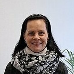 Frau steht lĂ€chelnd neben einem InformationsstĂ€nder mit BroschĂŒren und dem Logo âESOâ. Sie trĂ€gt einen schwarzen Pullover, eine helle Hose und einen gemusterten Schal. Im Hintergrund ist eine groĂe Fensterfront mit Ausblick auf ein BĂŒrogebĂ€ude und eine Topfpflanze.