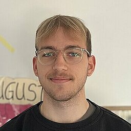 Ein junger Mann mit Brille, blondem Haar und kurzem Bart steht vor einer hellen Wand mit bunten Dekorationen, darunter die Monatsnamen âJuliâ und âAugustâ. Er trĂ€gt einen schwarzen Nike-Pullover und blickt freundlich in die Kamera. Im Hintergrund ist ein Teil eines Klassen- oder Gruppenraums zu erkennen.