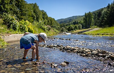 Ein kleiner Junge mit Kappe steht in einem Bergbach und bückt sich zum Wasser.