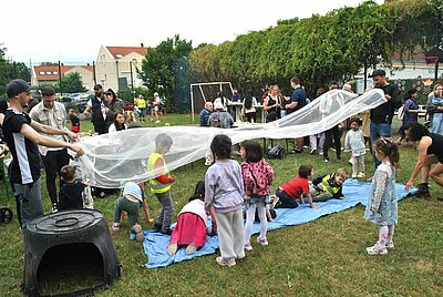 Sommerfest mit Spiel und Spaß Kinder krabbeln auf einer blauen Matte unter einem weißen Tuch, das von Erwachsenen über ihren Köpfen geschwungen wird. Im Hintergrund sitzen und stehen Eltern auf dem Außengelände der KITA.