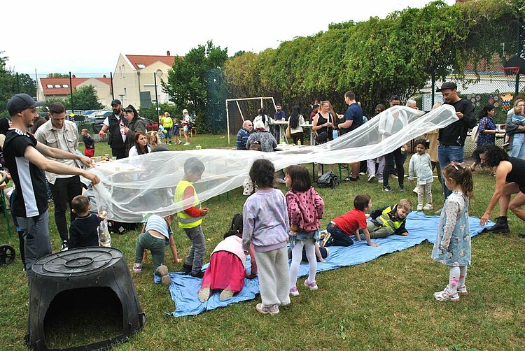 Sommerfest mit Spiel und Spaß Kinder krabbeln auf einer blauen Matte unter einem weißen Tuch, das von Erwachsenen über ihren Köpfen geschwungen wird. Im Hintergrund sitzen und stehen Eltern auf dem Außengelände der KITA.