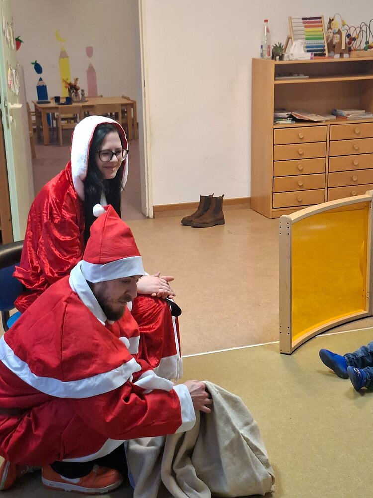 Eine als Weihnachtsfrau verkleidete Frau und ein als Weihnachtsmann verkleideter Mann sitzen lächelnd in einer KITA; der Mann hat einen Sack dabei und am rechten Bildrand sind die Füße eines Kindes zu sehen.