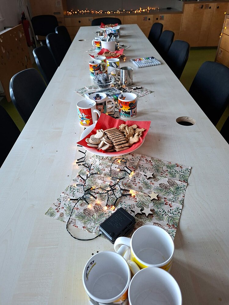 Langer Tisch mit vielen Stühlen, dekoriert mit Servietten im Tannenzapfenmuster, Lichterkette, Sternen, Kekstellern, Stiften und bunten Tassen – alles ist für das vorweihnachtliche Elterncafé vorbereitet.