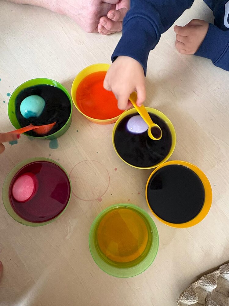 Schälchen mit verschiedenen Farben, darin liegen Eier zum Färben; an einer Schale ist eine Kinderhand zu sehen.