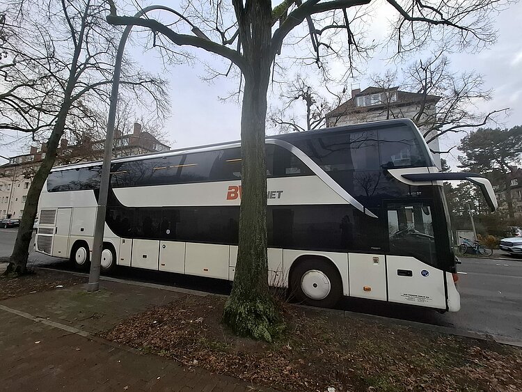 Ein großer weißer Reisebus parkt hinter zwei kahlen Bäumen.
