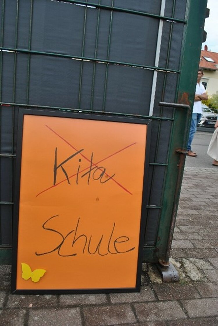 Großes Pappschild, auf dem das durchgestrichene Wort Kita und drunter Schule steht.
