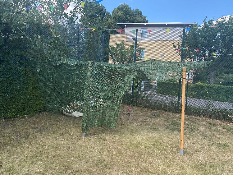Ein Gartenbereich mit einem Tarnnetz, das über einem Zaun und einem Teil des Rasens gespannt ist. Im Hintergrund sind ein Haus, Bäume und bunte Wimpelketten zu sehen.