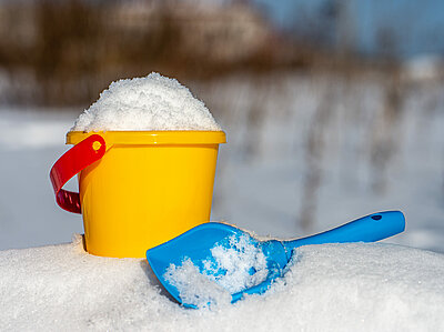 Ein gelber Eimer voller Schnee steht auf einem Schneehügel neben einer kleinen blauen Schaufel.